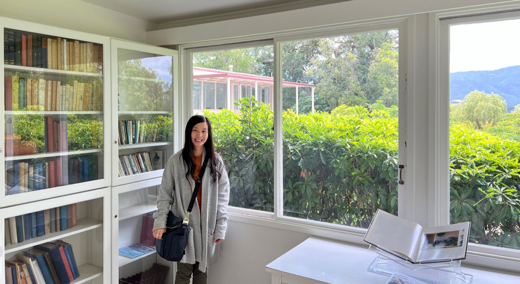 an image of Tiffany standing in a room with books and large windows, with greenery and mountains outside